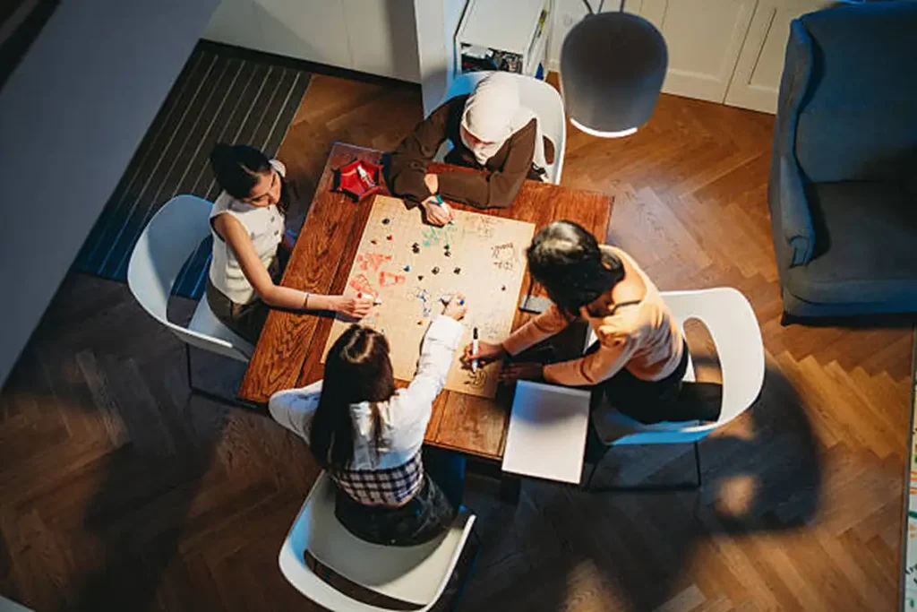 Vista aérea de quatro amigas sentadas ao redor de uma mesa de madeira, desenhando e jogando RPG de mesa em um mapa de papel.